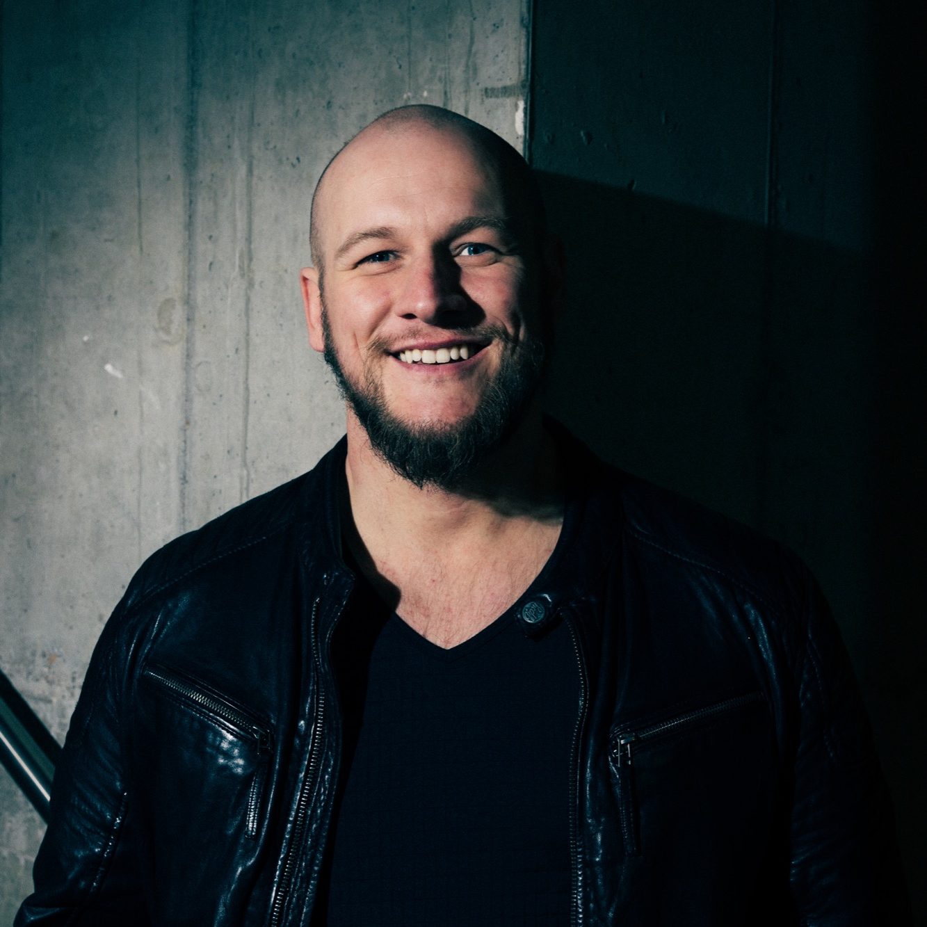 Tobias Mertens Musiker, Gitarrenlehrer, Schlagzeuglehrer in Hamburg Lächelnder Mann mit Glatze und Bart, trägt einen dunklen Hoodie.